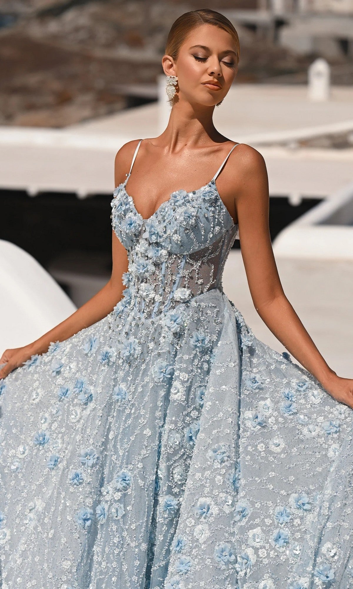 A woman in the Portia and Scarlett PS26027 long formal dress, featuring a light blue A-line silhouette with floral embellishments and thin straps, stands outdoors holding out the skirt of her gown.